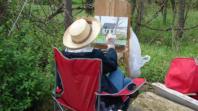 A woman seating in a red chair painting outside in a wooded area. 