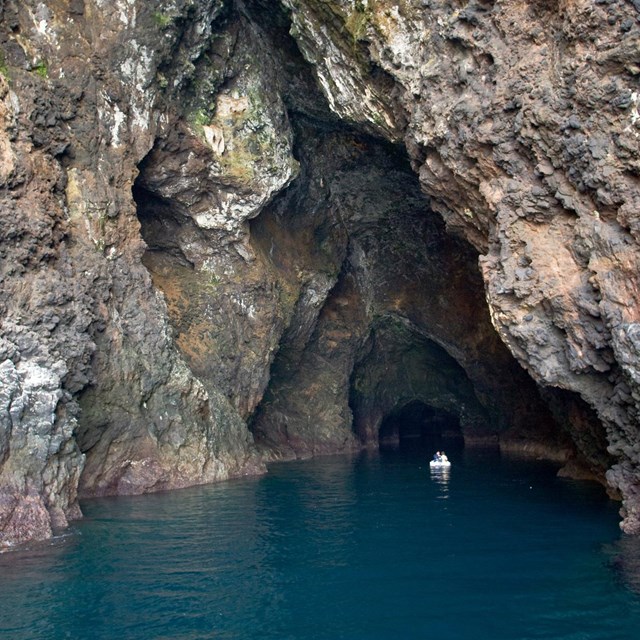 Sea cave with rust and black colored rocks and boat in the middle.