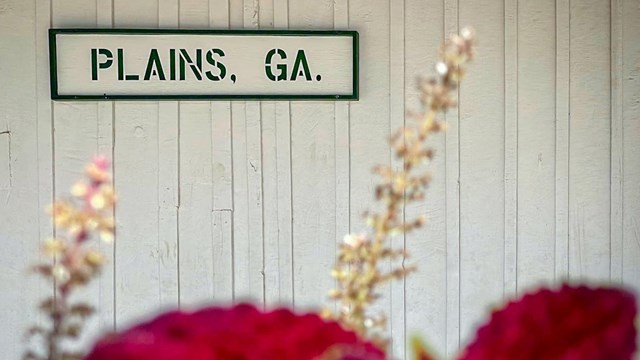 Plains, GA sign on wall at Plains Depot with flowers.
