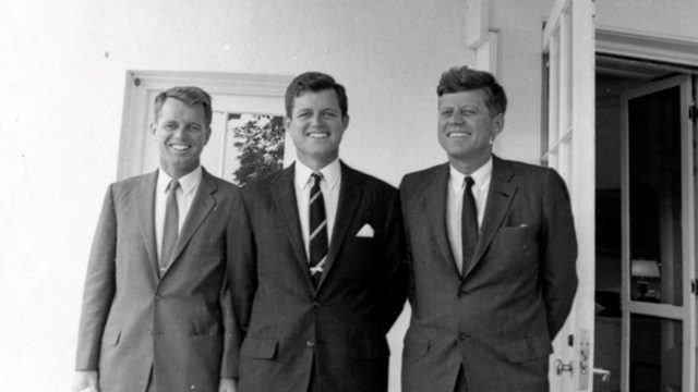 3 men in suits, Robert, Ted, and John F. Kennedy, stand outside the oval office.