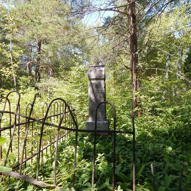 A grave marker is surrounded by a dilapidated fence in the forest.