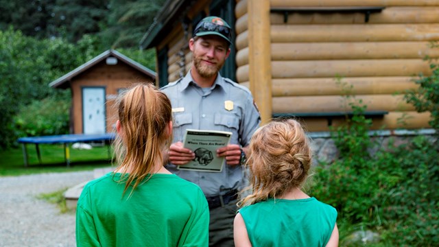 A ranger stands in front of two children in green shirts, holding a Junior Ranger Booklet.