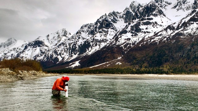 A person stands in a river looking through a research device with snowy mountains and clouds beyond.