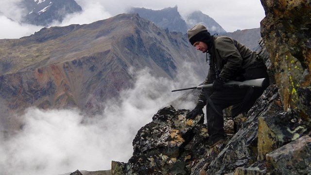 A man with a rifle sits on a steep, rocky slope