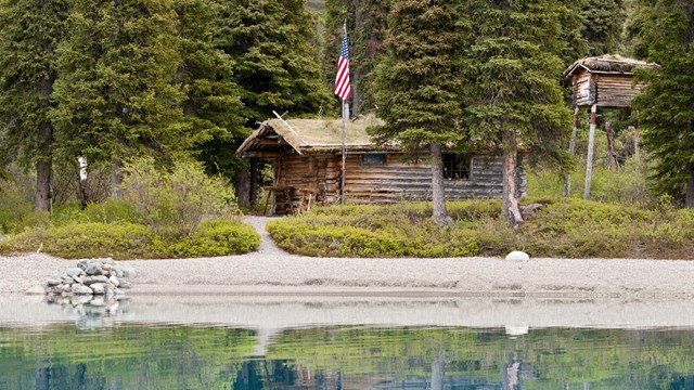 Calm blue water reflects a log cabin with a cache and American Flag surrounded by coniferous trees.