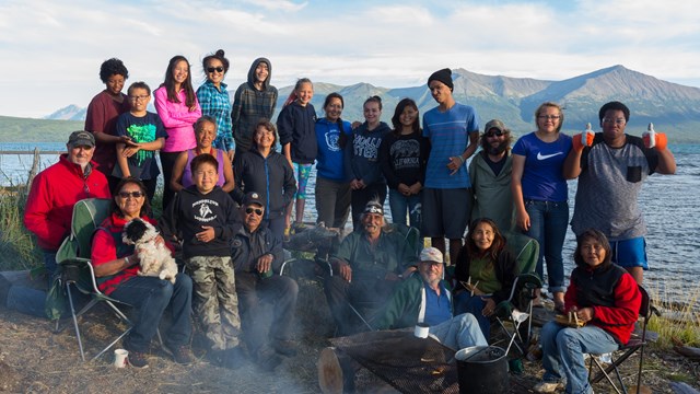 A group of people of all ages pose in front of a camp fire with water and mountains behind them.