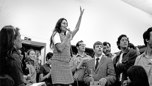 A woman stands on a chair surrounded by people while speaking into a microphone with her arm raised 