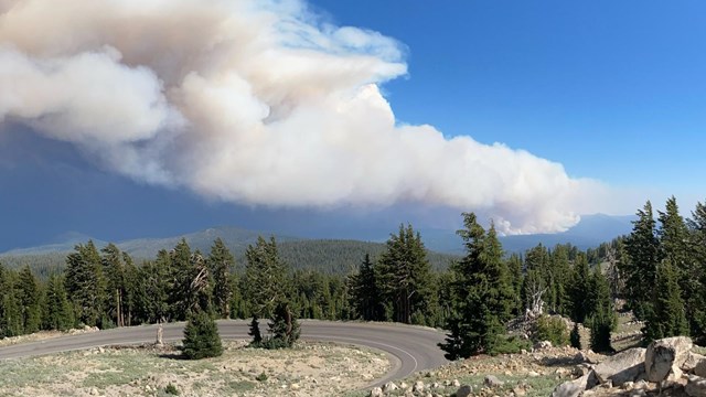 A road winds down a mountain slope with smoke rising from a wildfire in the distance. 