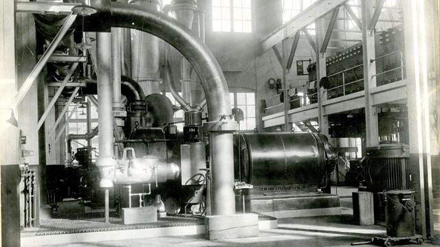 A steam engine and turbine are shown in a historic black and white photo, metal pipes crisscrossing