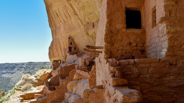 A stone masonry village and tower in a sandstone alcove with a canyon beyond