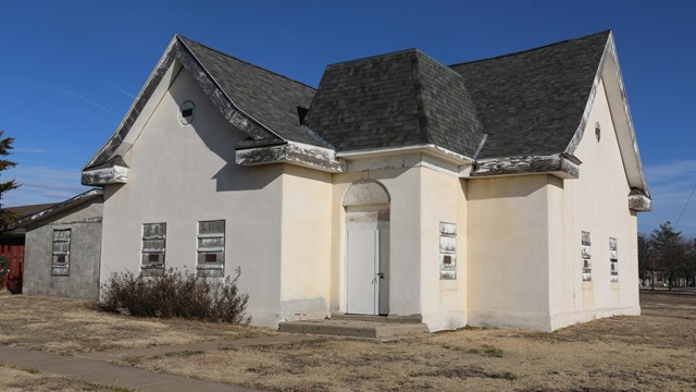 Side view of white stucco church building with gray shingles and scraggly rose bushes in front.