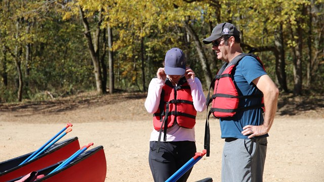 Two people stand wearing personal flotation devices, hats, and sunglasses.