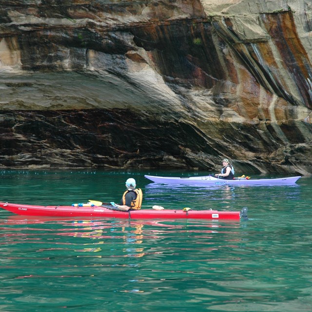 Two people in sea kayaks paddling along the colorful, dramatic Pictured Rock cliffs.
