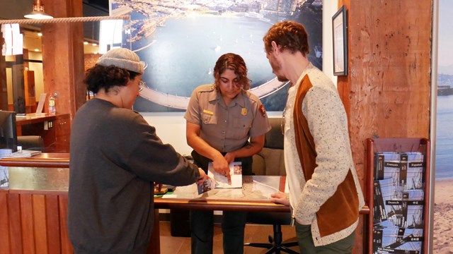 A park ranger at a visitor center desk points out directions on a map to two visitors