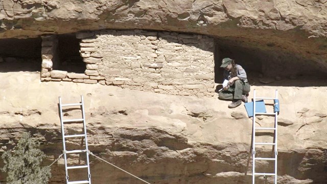 Person sitting beside a cliff dwelling recording notes.