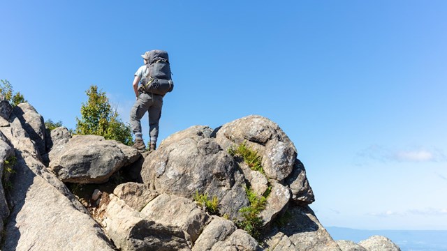 A man wearing a backpack stands on top of a series of boulders again a brilliant blue sky.