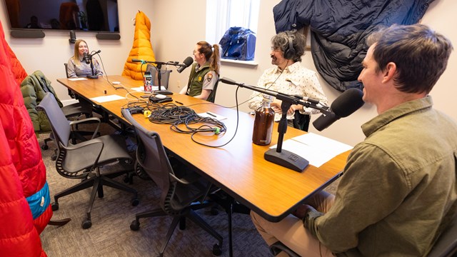 A group of park rangers, some in a green and grey uniform, sit around a table with microphones.
