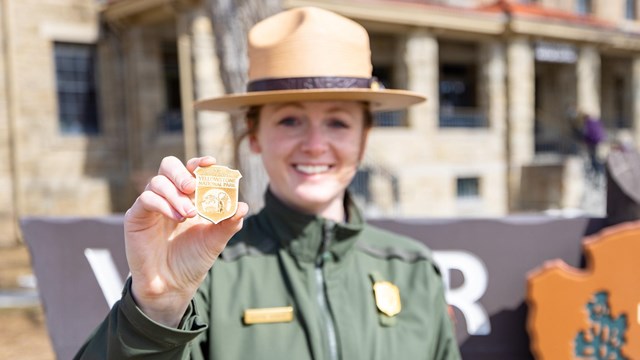 A Park Ranger holds up a wooden junior ranger badge in front of a visitor center.