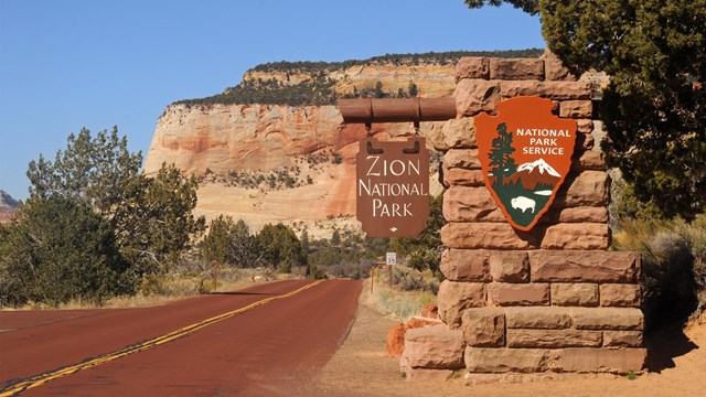 The Zion entrance sign, with a red road and red sandstone cliffs in the background. 