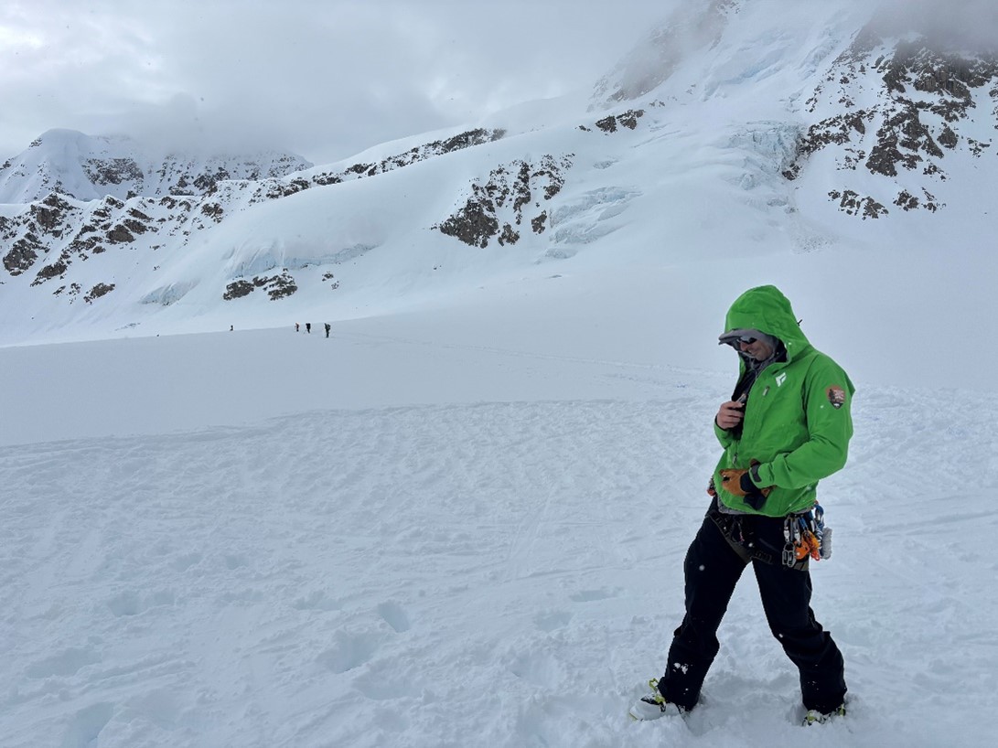 Rangers awaiting arrival of the NPS helicopter. Ranger in a green jacket standing on a snowy landscape with other rangers approaching in the distance.