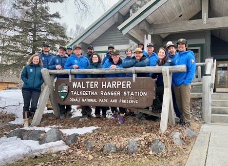 The 2026 Denali South District Team Eleven park rangers in blue jackets stand behind a sign that says Walter Harper Talkeetna Ranger Station Denali National Park and Preserve