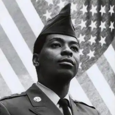 Black and white photo of a Black man wearing an Army uniform standing in front of an American flag.