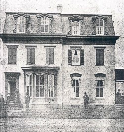 A black-and-white photograph of Frederick Douglass standing in front of a home
