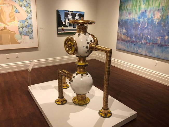 A large ceramic sculpture, with white spheres and brass metal looking pipes that resembles a piece of machinery.