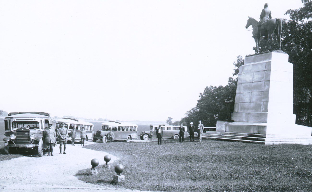 A black and white photograph of a tall white monument with a horse and rider on top is on the right. There is a curved dirt roadway on the left with four buses and their drivers parked and standing along the road.
