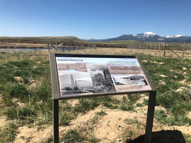 Interpretive wayside sign has historic image of smelter stack and large title "Pollution Stacks Up"