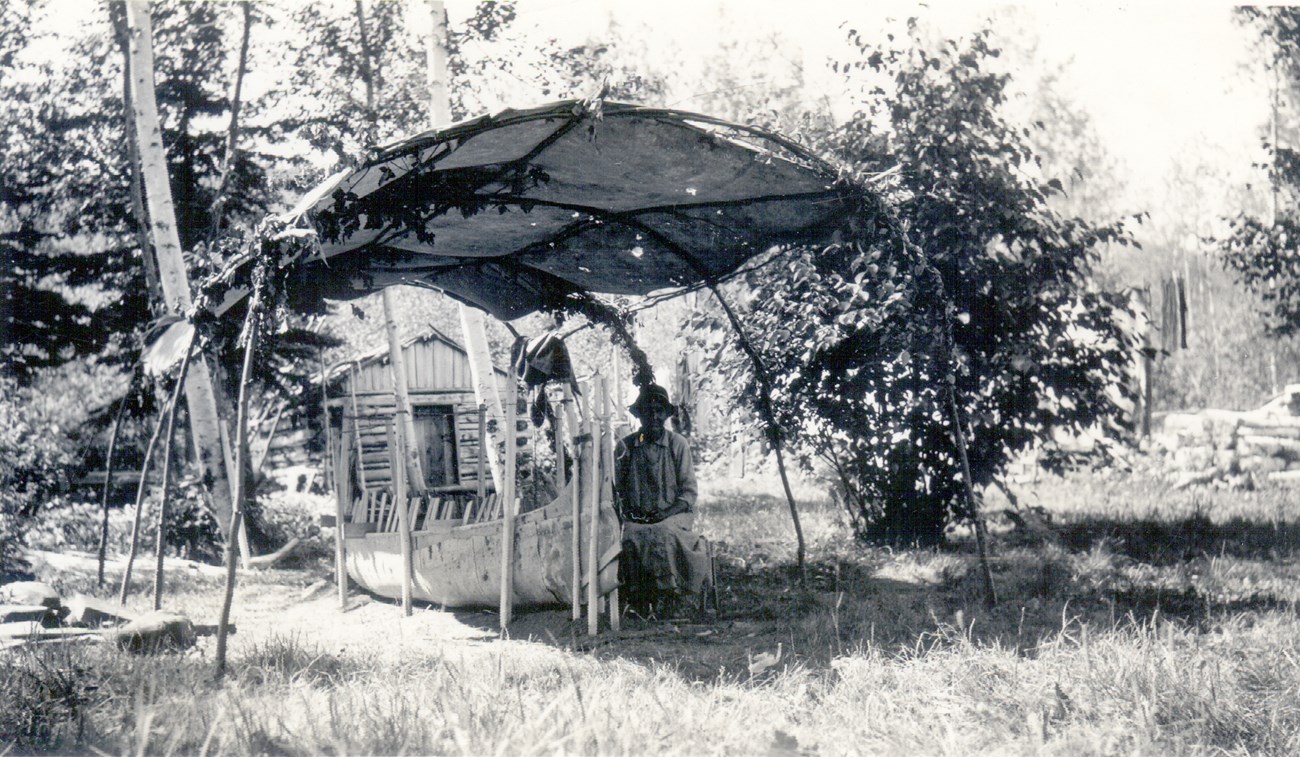 A person sitting next to a partially constructed birchbark canoe under a birchbark shelter.