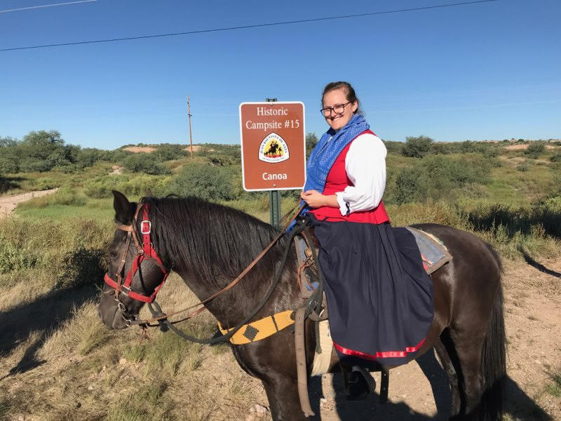 A woman dressed as an expedition member on horseback stands in front of a sign reading A woman dressed as an expedition member on horseback stands in front of a sign reading