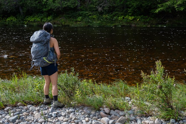 A hiker with a light grey backpack, navy shorts, and a tanktop stands on a shore of river rocks. She looks at the clear, light brown water of the large Wassataquoik Stream as it steadily flows in front of her.