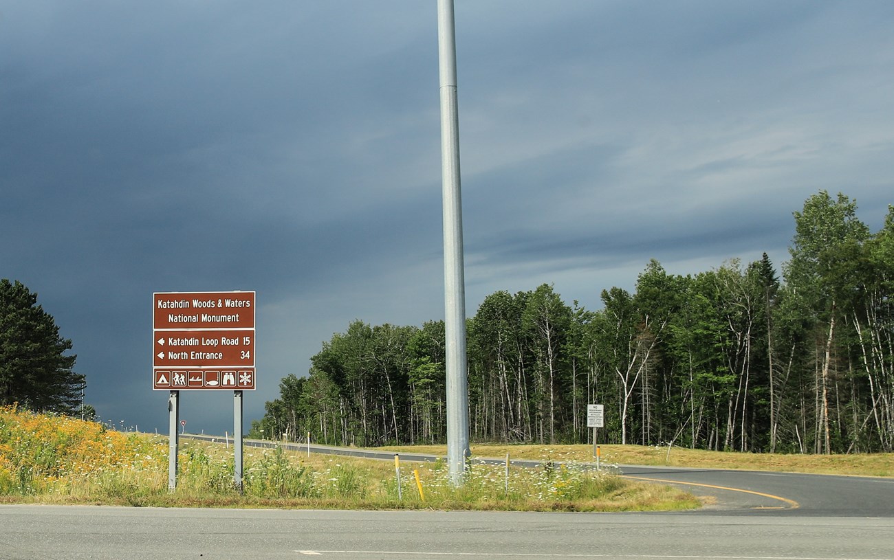 A colored photo of a large brown metal sign next to a road that shows directions to Katahdin Woods and Waters National Monument. The sign is next to a freeway on ramp.