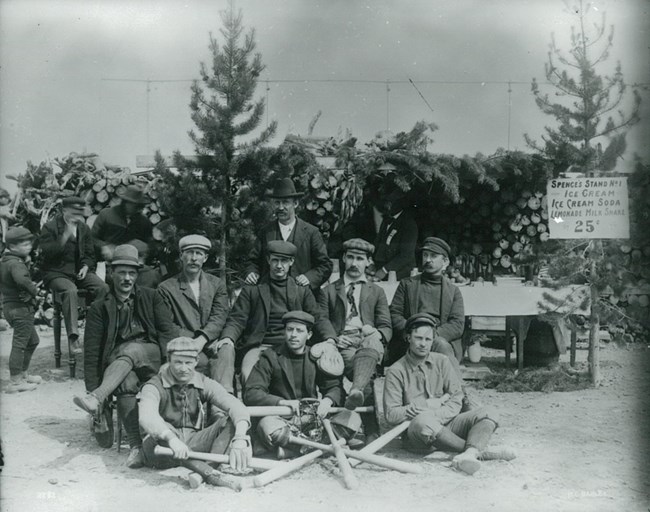 Black and white photo of nine men in three rows with baseball bats arranged in the foreground.
