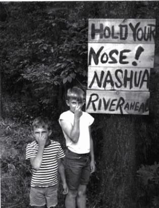 Two young boys are holding their noses next to a sign reading "Hold your nose! Nashua River ahead"