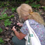 A blond woman kneels on the forest floor, breaking apart rich brown soil in her hands.