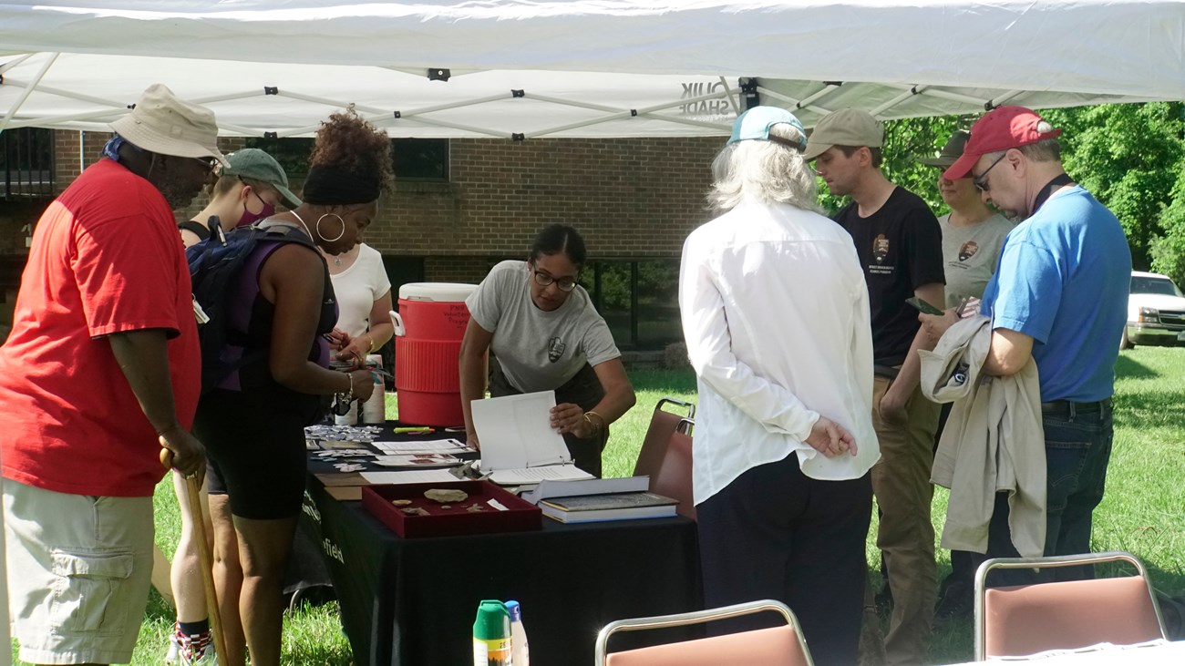 A black, female archeologist shows visitors artifacts from a recent dig at the Park. There are at a table under a shade tent.