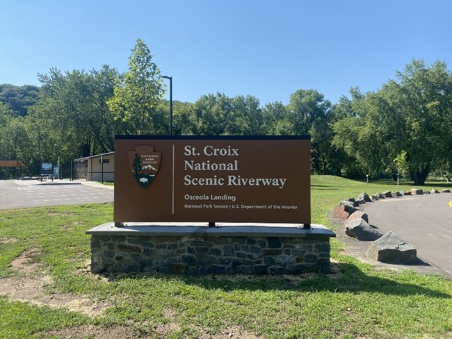 NPS sign in center of image with NPS arrowhead on the left and "St. Croix National Scenic Riverway Osceola Landing in white on the right."  The sign is attached at the bottom to a stone mount and is surrounded by grass with trees and parking lot behind.