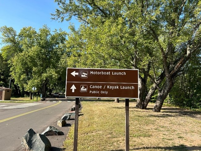 Brown sign in center with top half in white is an arrow pointing left, a motorboat icon, and "Motorboat Launch."  On the bottom half in white is an arrow pointing up, a canoe icon, and "Canoe Kayak Launch Public Only."  Sign has grass and trees around.