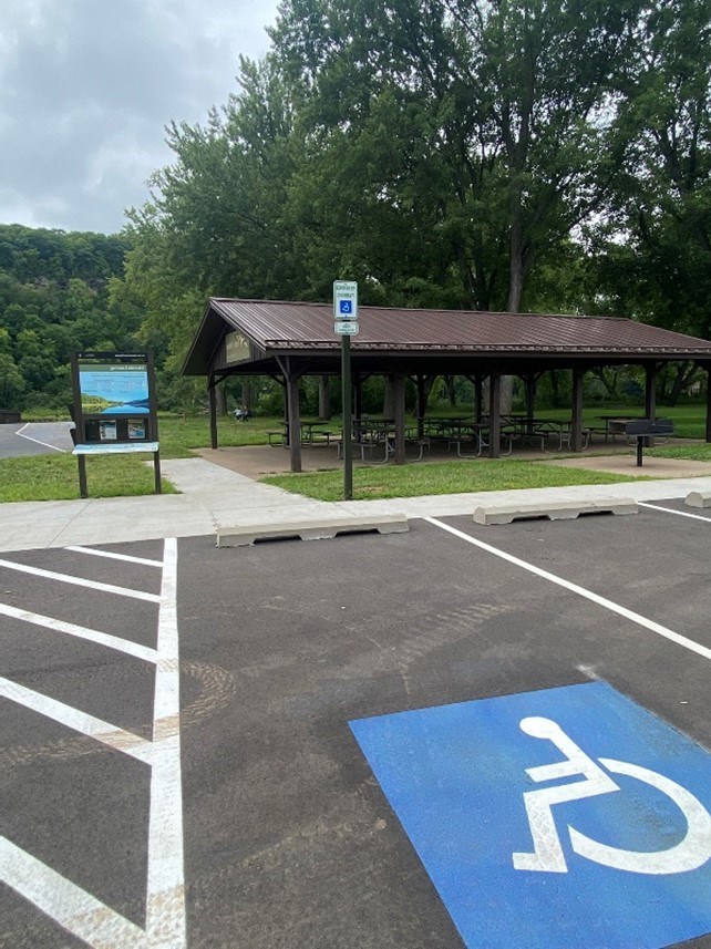 Accessible parking space in foreground with brown picnic shelter and blue orientation sign in mid image with green trees and light blue sky in background