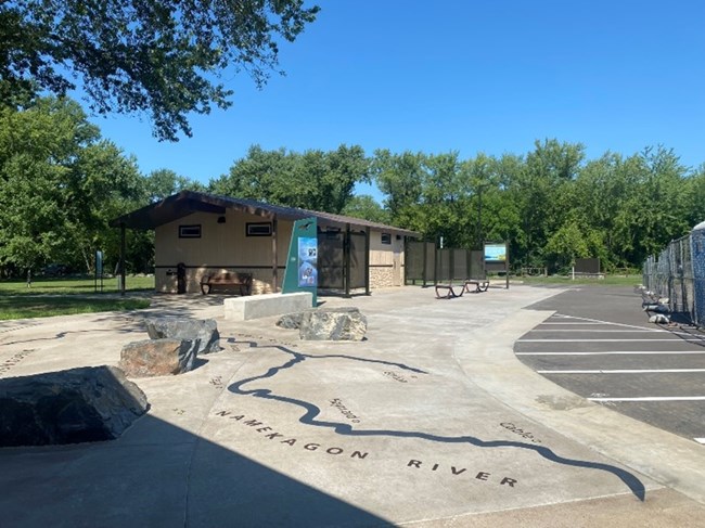 Grey concrete plaza in foreground with blue squiggly line and words "Namekagon River" etched into it.  Rocks and cement blocks for sitting and informational signs in mid image wiht tan and brown concrete building in background and parking lot on right