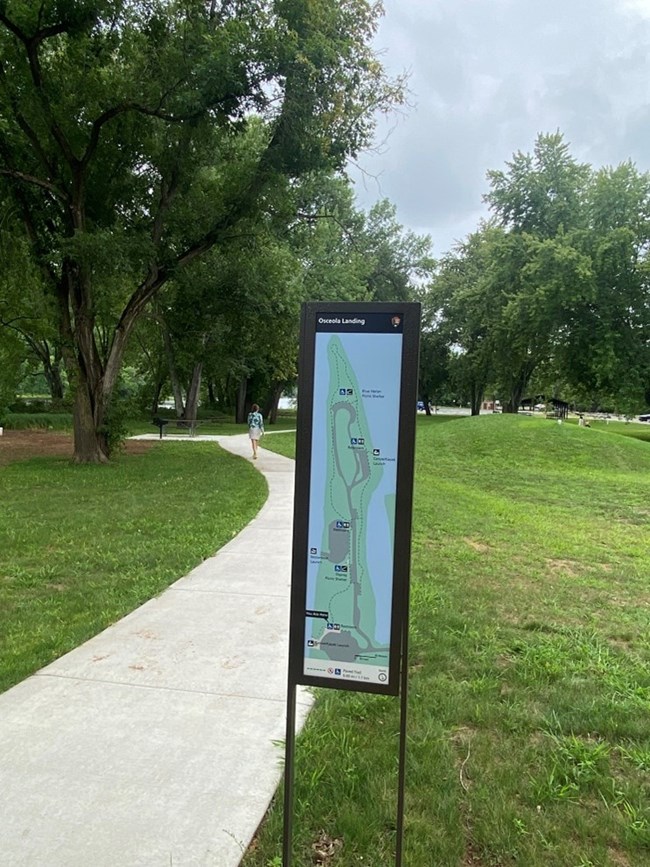 Tall, slim sign in center with outline of landing and roads.  Sign has a concrete path on the left, grass on the right, and trees in the background.