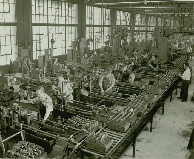 Rows of heavy machinery by a brick wall with large windows. Men are busy at work at the machines.