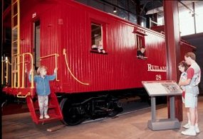 parents read an exhibit panel as a little girl comes down the steps of a red caboose in Steamtown's Technology Museum