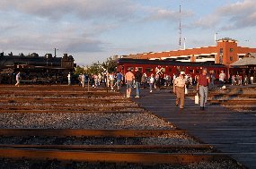 visitors walk accross railroad tracks with a steam locomotive and red passenger car sitting by a loading platform in the background visitors walk accross railroad tracks with a steam locomotive and red passenger car sitting by a loading platform in the background