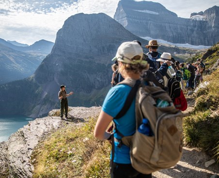 Visitors on trail listen to ranger standing on cliff edge above a mountain valley with lake