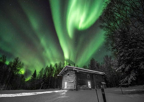 Green lights swirl in the night sky over a wooden cabin in snowy woods.
