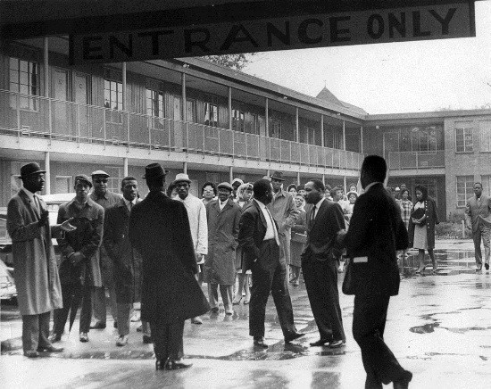 A group of men in overcoats gather in a rainy courtyard of a two-story motel.
