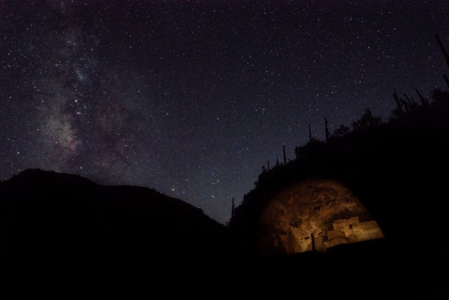 An illuminated cliff dwelling with the Milky Way galaxy above it.